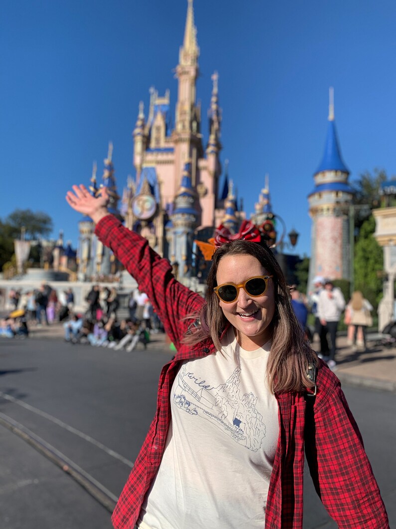 May include: A person wearing a white t-shirt with a mountain graphic and the word "wild" in blue, paired with a red and black plaid shirt and yellow sunglasses. The background features a large castle under a clear blue sky.