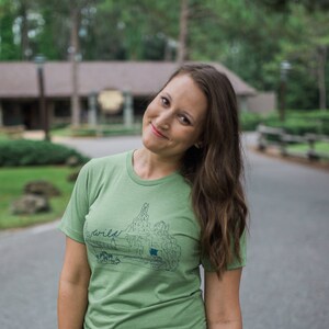 May include: A light green t-shirt with a graphic of a landscape and the word "wild" in a handwritten font. The shirt is being worn by a person with long brown hair.