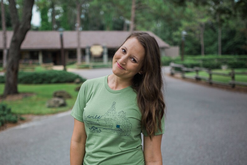 May include: A light green t-shirt with a graphic of a landscape and the word "wild" in a handwritten font. The shirt is being worn by a person with long brown hair.
