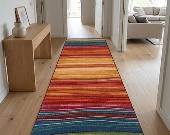 May include: A long rectangular runner rug featuring a horizontal striped pattern with a vibrant gradient of colours including red, orange, yellow, blue, and green. The rug has a thin dark red border and a textured appearance throughout the surface.