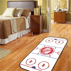 Könnte beinhalten: Ein Teppich im Hockey-Rink-Stil mit dem Logo der Detroit Red Wings in der Mitte. Der Teppich hat einen weißen Hintergrund mit roten und blauen Linien, Kreisen und Hash-Markierungen, die einem Hockey-Rink nachempfunden sind. Der Teppich liegt auf einem Holzboden.