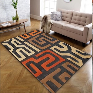 May include: A rectangular rug featuring a geometric design in black, orange, brown, and beige. The rug is laid on a wooden floor. A beige sofa is visible in the background, with a fur throw.