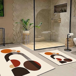 Op de afbeelding: Twee abstracte geometrische badmatten in beige, bruin, oranje en rood. De matten liggen op een betegelde vloer in een badkamer.
