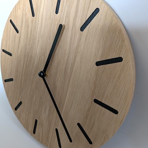 May include: A round wooden wall clock with a light brown finish. The clock features black hour markers and black clock hands. The clock is mounted on a white wall. The clock's diameter is approximately 12 inches.