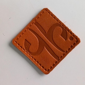 May include: A brown leather label with a stitched border and a raised embossed design. The design features a stylized letter "C" with a curved shape.