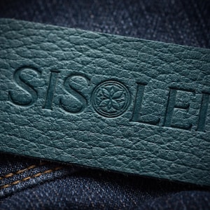 May include: Close-up of a teal leather label with the embossed word "SISOLEI" and a floral emblem. The label is rectangular and has a textured surface. The label is set against a dark blue denim background.