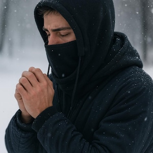 May include: A dark navy blue hooded jacket with a built-in face covering. The person is wearing the hood and face covering, with their hands clasped together. Snowflakes are falling in the background.