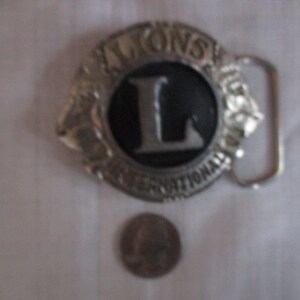Puede incluir: Hebilla de cintur&oacute;n de plata con un dise&ntilde;o en blanco y negro. La hebilla tiene el texto "LIONS INTERNATIONAL" y una gran letra "L" en el centro.