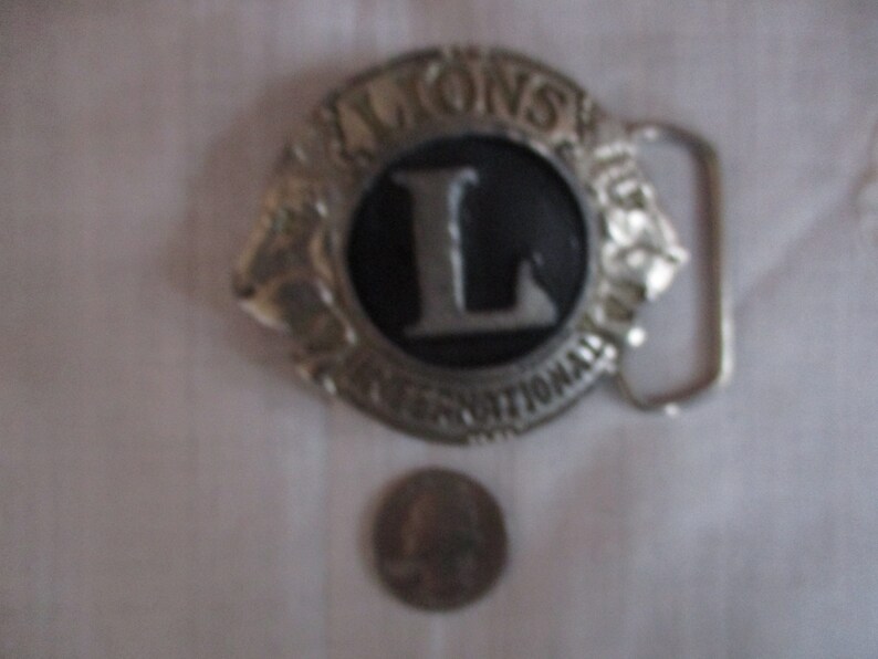 Puede incluir: Hebilla de cintur&oacute;n de plata con un dise&ntilde;o en blanco y negro. La hebilla tiene el texto "LIONS INTERNATIONAL" y una gran letra "L" en el centro.