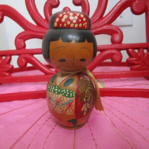 May include: A wooden Japanese kokeshi doll with a red and white patterned hat, black hair, and a colorful kimono. The doll is painted with a floral design and has a red sash.