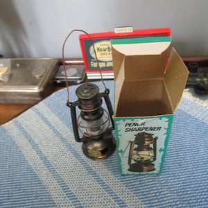 May include: Miniature lantern pencil sharpener with a clear glass globe and black metal frame. The lantern sits next to its box, which features a matching lantern illustration and the words "Pencil Sharpener". The box is teal and brown.