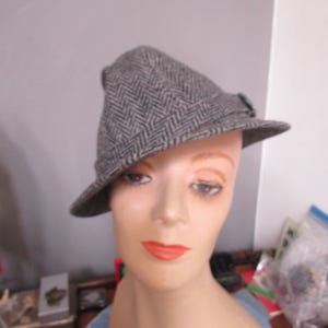 May include: A gray herringbone tweed hat with a small brim and a decorative buckle. The hat is displayed on a mannequin head with red lipstick. The hat is a classic style.