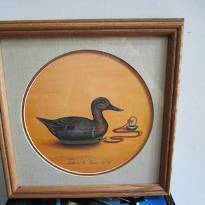 May include: Framed print of a painting of a duck with a chain and a small object. The painting is on a yellow background and is signed "David T. Klein" in the bottom right corner.