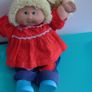 May include: A vintage doll with a soft, vinyl face and light blonde yarn hair styled in a ponytail. The doll wears a red polka-dot dress with white lace trim, blue denim pants, and light blue shoes. The doll's eyes are blue.