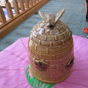 May include: A ceramic honey pot shaped like a beehive with three bees on the bottom and a bee on the top. The beehive is brown and yellow with a textured surface.