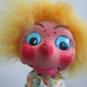 May include: A pink puppet with a large nose, blue eyes, and yellow hair. The puppet is wearing a floral dress.