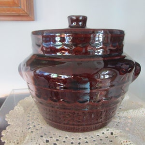 May include: A brown glazed ceramic crock with a lid. The crock has a textured surface and a rounded shape. The lid has a knob on top.