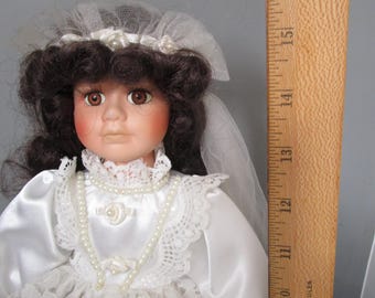 May include: A porcelain doll wearing a white satin and lace dress with a pearl necklace. The doll has brown hair, brown eyes, and rosy cheeks. The doll is approximately 14 inches tall.