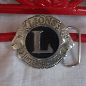 Puede incluir: Hebilla de cintur&oacute;n de plata con un logotipo de Leones Internacional en blanco y negro con una gran letra L. La hebilla tiene un dise&ntilde;o de cabeza de le&oacute;n a cada lado.