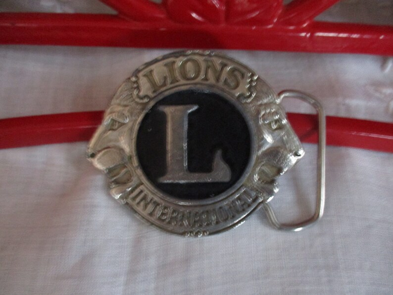 Puede incluir: Hebilla de cintur&oacute;n de plata con un logotipo de Leones Internacional en blanco y negro con una gran letra L. La hebilla tiene un dise&ntilde;o de cabeza de le&oacute;n a cada lado.