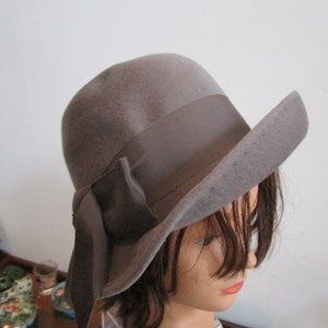 May include: A gray felt fedora hat with a wide brim and a large gray satin bow. The hat is sitting on a mannequin head with short brown hair.