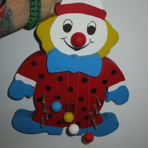 May include: A wooden clown toy with a red and white hat, a yellow face, and a blue bow tie. The clown has black polka dots on its red shirt and is holding a string with colorful balls.