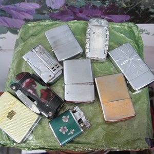 May include: A collection of vintage lighters in various styles and colors, including silver, gold, and green. Some lighters have intricate designs, while others are plain. The lighters are arranged on a green surface.
