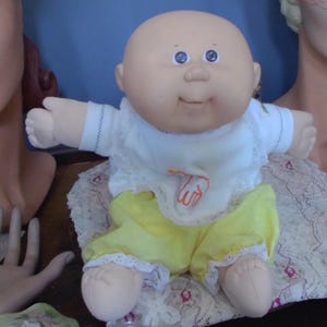 May include: A vintage Cabbage Patch Kid doll with a bald head, blue eyes, and a smiling expression. The doll wears a white shirt with a lace collar and yellow shorts. The doll is sitting on a patterned surface.