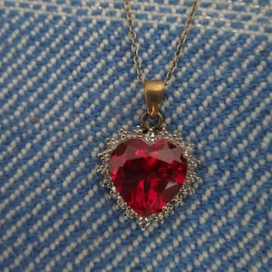 May include: A silver necklace with a heart-shaped pendant. The pendant features a large, faceted red gemstone surrounded by a halo of smaller, clear stones. The necklace hangs against a blue and white patterned background.