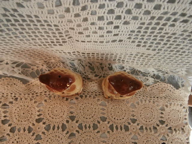 May include: A pair of ceramic hedgehog-shaped salt and pepper shakers. The hedgehogs have brown snouts and backs, and cream-coloured bodies. Each shaker has two holes for dispensing salt or pepper. They are placed on a beige crocheted surface.