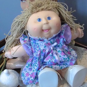 May include: A vintage Cabbage Patch Kid doll with blonde yarn hair, blue eyes, and rosy cheeks. The doll wears a floral print dress with a purple collar and sleeves. White shoes with laces complete the look. A collectible doll.