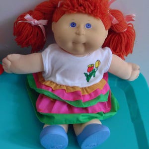 May include: A vintage doll with bright orange yarn hair styled in pigtails, tied with pink ribbons. The doll has blue eyes, a white shirt with a tulip design, and a layered skirt in pink, orange, and green. Blue shoes complete the look.