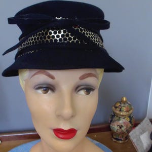 May include: A dark navy blue felt hat featuring a structured crown and a slightly flared brim. The hat is accented with a decorative band of metallic gold material with a repeating circular cutout pattern and a tied bow detail at the front.
