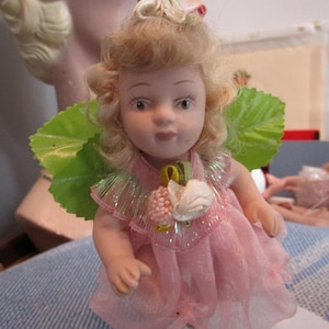 May include: A miniature doll with blonde curly hair, wearing a pink dress with a sheer skirt and a white swan embellishment. The doll has green leaf-shaped wings and is suspended by a gold string, suggesting it's an ornament.