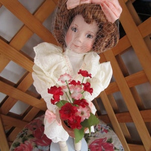 May include: A porcelain doll with a white dress and a pink bow in her hair is holding a pot of red and pink flowers.