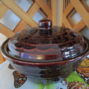 May include: A brown glazed ceramic casserole dish with a lid. The dish has a rounded shape and a glossy finish.