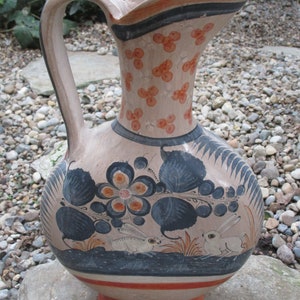 May include: A hand-painted ceramic pitcher with a wide spout and a handle. The pitcher is decorated with blue and white floral designs, orange accents, and two white rabbits. The pitcher is sitting on a gray stone surface.