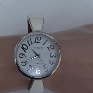 May include: Silver-toned Studio quartz watch with a white face and black numerals. The watch has a silver band and is displayed on a light-colored surface. The watch face reads "Studio!" and "Quartz".