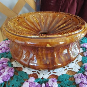 May include: A brown glazed ceramic pedestal with a fluted top and a wide base. The pedestal is sitting on a crocheted doily with purple and green accents.