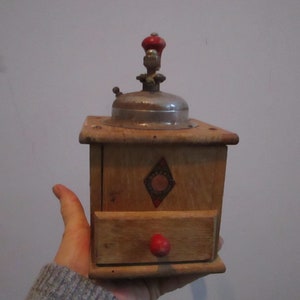 May include: A vintage wooden coffee grinder with a metal top and a red knob. The grinder has a drawer for collecting the ground coffee.