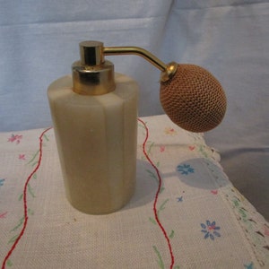 May include: A white and gold atomizer bottle with a brown fabric atomizer top. The bottle is sitting on a white embroidered tablecloth.