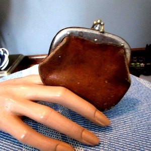 May include: A vintage brown leather coin purse with a metal clasp. The purse has a rounded shape and a worn appearance, suggesting age and use. The clasp is silver-toned, and the purse is resting on a blue and white striped surface.