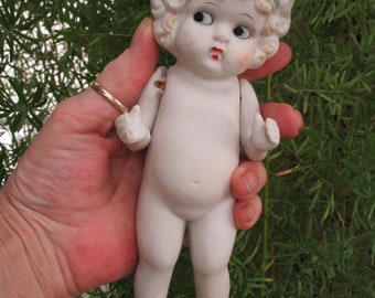 May include: A porcelain doll with a white dress and a pink flower on her hat. The doll has blonde hair and blue eyes.