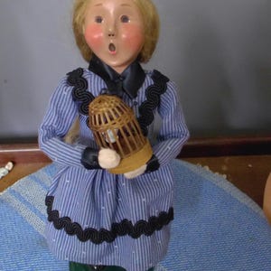May include: A vintage figurine of a woman with blonde hair and rosy cheeks, wearing a blue striped dress with black trim. She holds a small, brown birdcage. The figurine is mounted on a green base.