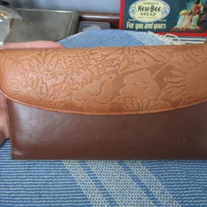 May include: A brown leather wallet with a floral embossed design on the flap. The wallet has a bi-fold design with a darker brown base and a lighter brown flap. The wallet is a classic accessory for carrying cards and cash.