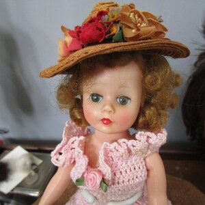 May include: A vintage doll with curly blonde hair wearing a pink crocheted dress and a straw hat with floral accents.