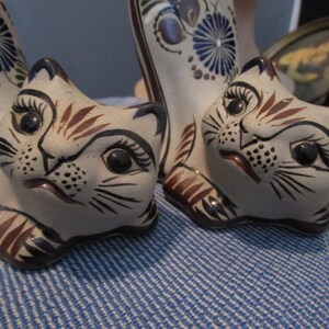 May include: Hand-painted ceramic cat figurines. The cats have a cream-colored base with brown and black details. The eyes are large and black, and the whiskers are finely drawn. The figurines are on a blue and white striped surface.