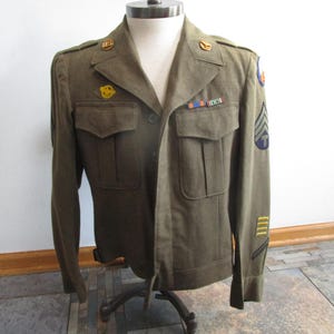 May include: Olive green military jacket with multiple pockets, buttons, and patches. The jacket features insignia on the shoulders and sleeves, including stripes and a circular patch. The jacket is displayed on a mannequin.