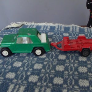May include: A vintage green toy car with a white interior and white wheels is connected to a red trailer. The toy car has a convertible top. The toy car and trailer are on a blue and white patterned surface.