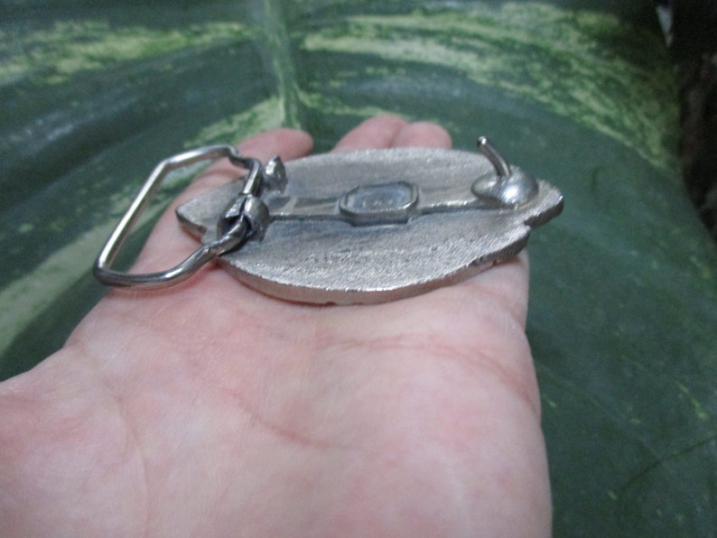 Puede incluir: Hebilla de cintur&oacute;n plateada con un dise&ntilde;o detallado de una hoja. La hebilla tiene un cierre de gancho y un acabado liso y pulido.